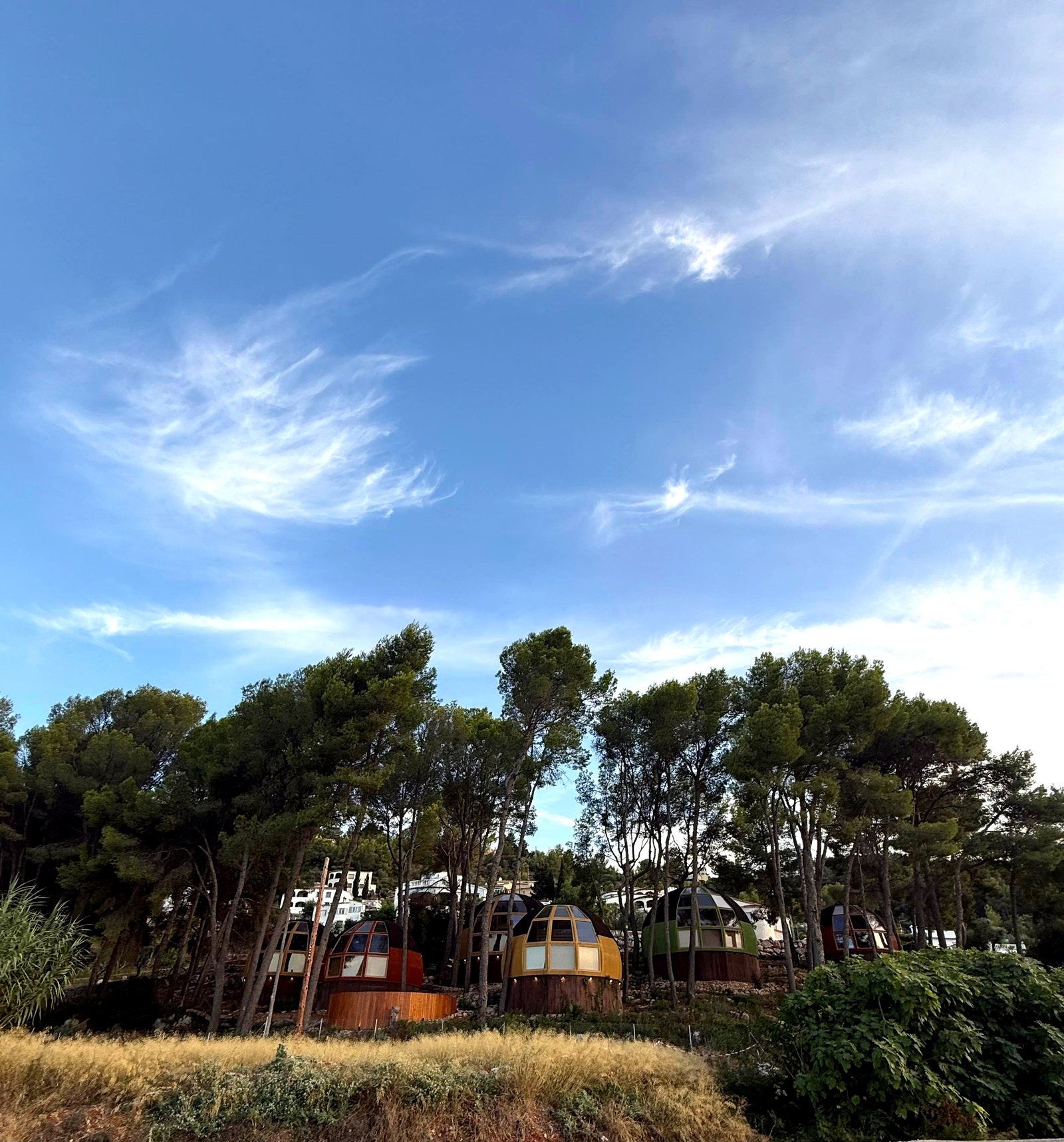 Las seis casas domo geodésicas de AUMARA en colores naranja, amarillo, verde y rojo bajo un amplio cielo azul en Benidoleig, Alicante (Costa Blanca)