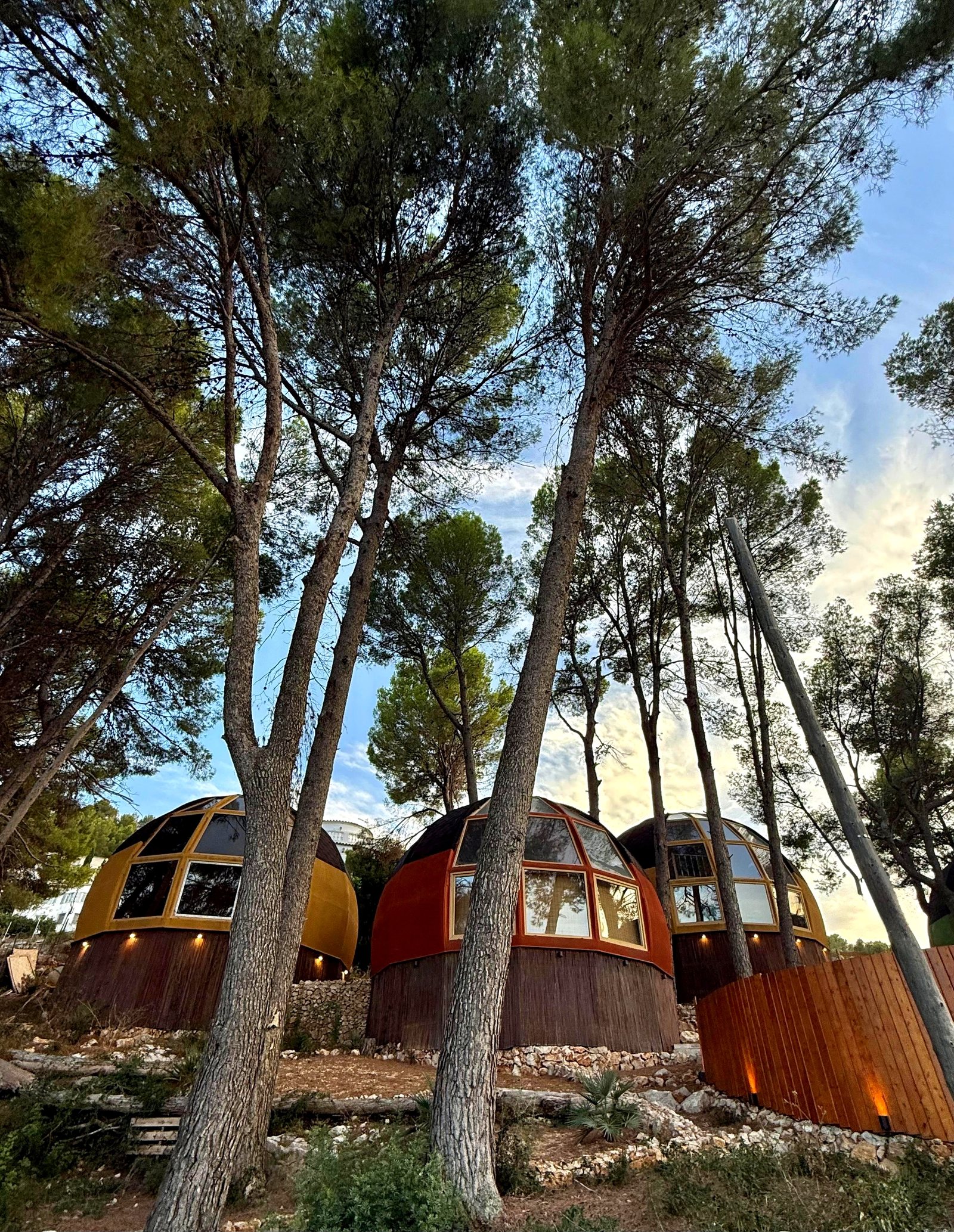 Casa domo geodésica naranja de AUMARA con ventanales, entre un domo amarillo y otro ocre, con valla de madera iluminada en Benidoleig, Alicante (Costa Blanca)